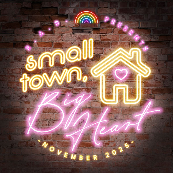 A neon sign on a dark brick wall. The sign says “Small Town, Big Heart” in yellow and pink neon text. The yellow outline of a house with a pink heart inside accompanies the text. Pink text is curved around the top and says "R. A. D. Presents.". A neon rainbow glows in between RAD and Presents. Text is along the bottom and says "- November 2025 -" in white neon font.